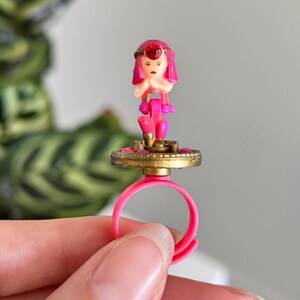 May include: A vintage pink plastic ring with a small figurine on top. The figurine has a pink dress and a red gem on its head. The ring's base is gold-colored and circular.