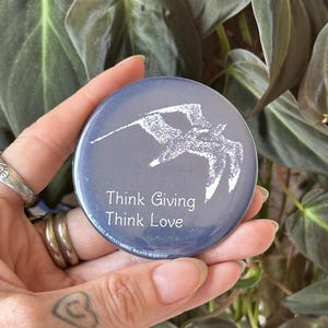 Puede incluir: Una chapa redonda azul con un gráfico blanco de un pájaro en vuelo. El texto "Think Giving Think Love" está impreso en blanco debajo del pájaro, con "1973 HALL BARTLETT ALL RIGHTS RESERVED" escrito a lo largo del borde inferior.