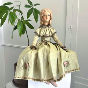 May include: A vintage doll in a light green dress with a ruffled collar and long sleeves. The dress features embroidered pink roses and a dark ribbon. The doll has a painted face, blonde hair, and is seated.