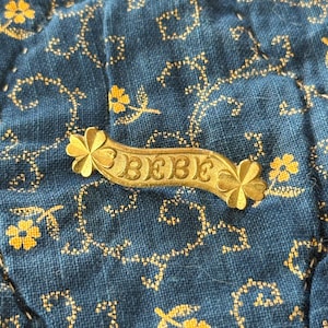 K&ouml;nnte beinhalten: Eine goldfarbene Metall-Anstecknadel mit dem Wort "BEBE" in der Mitte. Die Nadel hat an jedem Ende ein Blumendesign. Die Nadel befindet sich auf einem blauen Stoffhintergrund mit einem floralen Muster.