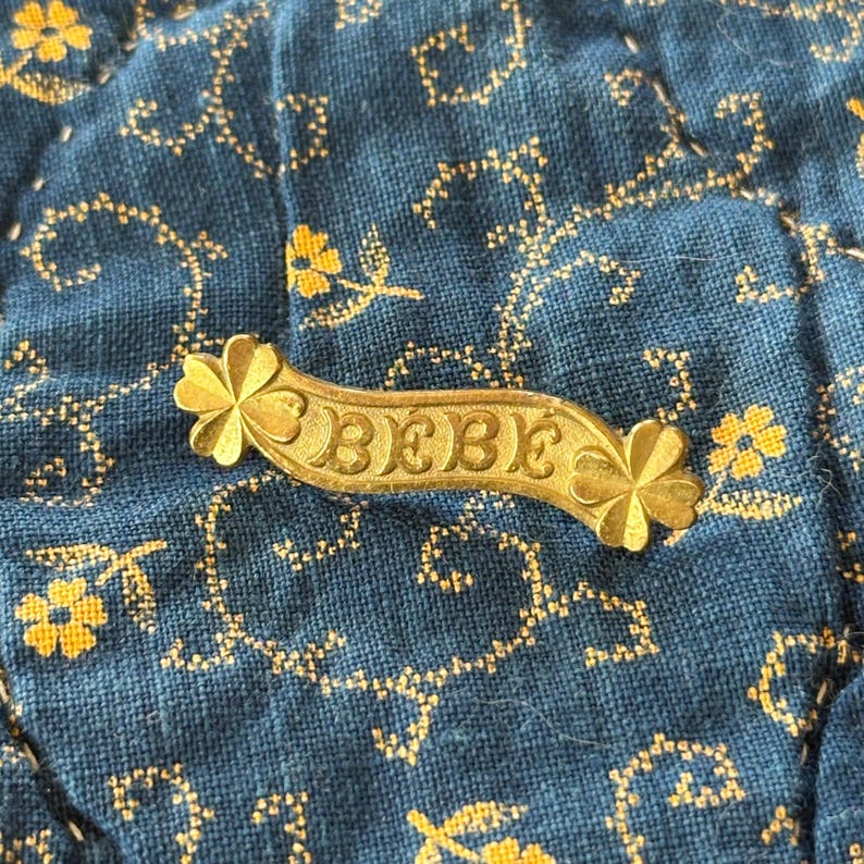 K&ouml;nnte beinhalten: Eine goldfarbene Metall-Anstecknadel mit dem Wort "BEBE" in der Mitte. Die Nadel hat an jedem Ende ein Blumendesign. Die Nadel befindet sich auf einem blauen Stoffhintergrund mit einem floralen Muster.