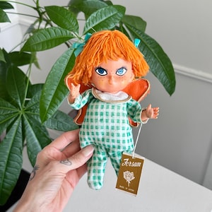 Puede incluir: Una muñeca vintage con cabello de hilo naranja, ojos azules y alas naranjas. La muñeca viste un atuendo a cuadros verdes y blancos. Una etiqueta dice "Forsum" y "Made in Japan".