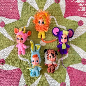 May include: Five small dolls dressed in animal costumes. One wears a pink horned hood, one has a bright orange lion mane, one is a blue rabbit with long ears, one is a tiger with black stripes, and one is a purple mouse with large round ears.