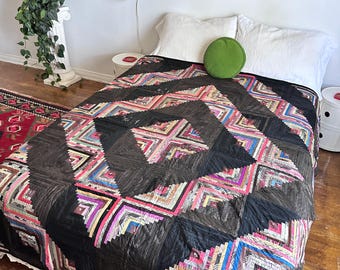 Pode incluir: Uma colcha com padrão de cabana de troncos, apresentando uma grande forma de diamante preta central rodeada por bordas geométricas concêntricas em tons de rosa, azul, amarelo e branco, sobre uma cama com duas almofadas brancas e uma almofada verde.
