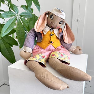 May include: A vintage stuffed rabbit doll with long ears, wearing a purple jacket, yellow vest, pink pants, and a white cap. The doll has painted facial features and is seated on a white surface.
