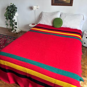 May include: A large red wool blanket featuring horizontal stripes in black, yellow, and green across the top and bottom edges. The blanket is draped over a bed with two white pillows and a small, round, solid green decorative throw pillow.