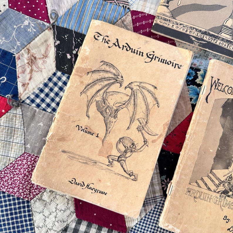 Puede incluir: Libro vintage "The Arduin Grimoire" Volumen 1 con una ilustraci&oacute;n fant&aacute;stica de una criatura alada y una figura con un arma. El libro es de color beige con texto e ilustraciones en negro. El libro est&aacute; sobre un edred&oacute;n de retazos.