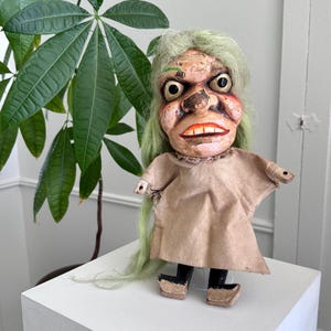May include: A vintage folk art doll with a grotesque face, green hair, and a tan paper dress. The doll has black shoes and is standing on a white surface. A large green plant is in the background.