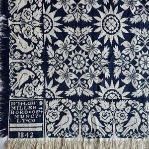 May include: A navy blue and white woven textile featuring a repeating floral and bird pattern. The fabric includes a rectangular block with the text WM. LOW MILLER BORO OF MUNCY LY. CO and the date 1843. The edges are finished with a fringe trim.