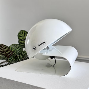 May include: A white, mid-century modern desk lamp with a curved base and a domed shade. The lamp has a minimalist design and features the text "iGuzzini made in Italy". The lamp is on a white surface.
