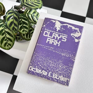 May include: A purple paperback book titled "Clay's Ark" by Octavia E. Butler. The book cover features a blurry image of a world map and the text "A Novel of Science Fiction".