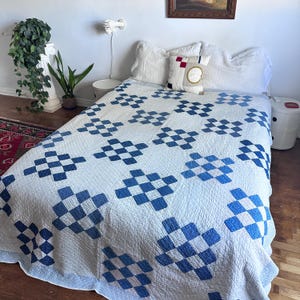 May include: A white and blue quilt with a repeating pattern of blue squares. The quilt is on a bed with white pillows and a decorative pillow. The room has a vintage aesthetic.