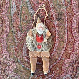 Può includere: Un ornamento da appendere a forma di figura con volto barbuto bianco, cappello a punta color marrone chiaro e braccia e gambe in tessuto dello stesso colore. Il busto è una sacca a rete color marrone chiaro con una mezzaluna rossa.