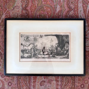May include: A black and white etching print in a simple black frame with a wide off-white mat. The artwork depicts a detailed interior scene with multiple figures, including a person playing an accordion, various objects, and architectural elements.