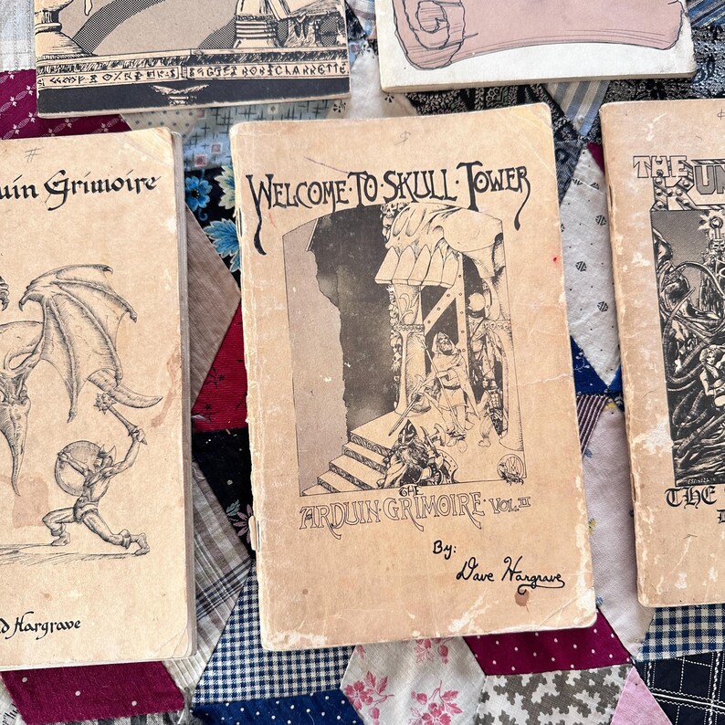 Puede incluir: Una colecci&oacute;n de libros antiguos con cubiertas de color beige envejecido. El libro central presenta el t&iacute;tulo "WELCOME TO SKULL TOWER" en una fuente g&oacute;tica, con ilustraciones de figuras y una torre. Otros libros incluyen "Arduin Grimoire".