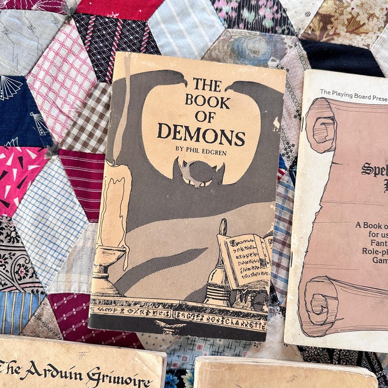 Puede incluir: Libro vintage titulado "The Book of Demons" de Phil Edgren, con una silueta de murci&eacute;lago e ilustraci&oacute;n de vela. Se ven otros libros, incluido uno titulado "Spells". Los libros est&aacute;n sobre un edred&oacute;n de retazos.