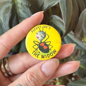 May include: A round yellow pinback button featuring a cartoon spider with long eyelashes and red lips. The text "SLEEPY JOE'S" is printed in black above the spider, and "THE WIDOW" is printed in black below the spider, which has a red hourglass mark.