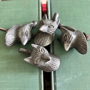 May include: Four antique-style cabinet knobs with fox head designs. The knobs are made of a dark, metallic material and feature detailed fox faces. The knobs are arranged on a green surface, suggesting a decorative or hardware theme.