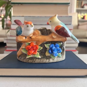 May include: A ceramic bird figurine featuring two colorful birds perched on a brown tree branch. The base is decorated with red and blue flowers. The birds have orange, blue, and white details. A decorative item for home decor.