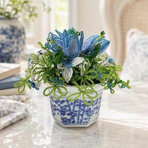 Könnte beinhalten: Ein dekoratives Blumenarrangement aus kleinen blauen, weißen und grünen Glasperlen. Die perlenbesetzten Blumen und Blätter befinden sich in einem weißen, sechseckigen Keramiktopf, der außen mit einem blauen Bambusmuster bemalt ist.