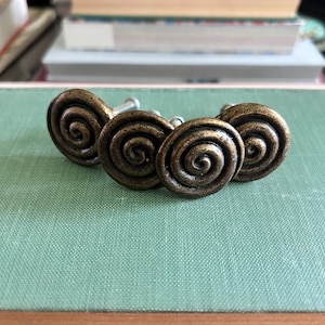 Puede incluir: Cuatro tiradores de armario antiguos de color bronce con un diseño en espiral. Los tiradores están dispuestos en fila sobre una superficie verde, con libros en el fondo. Cada tirador tiene un diámetro de aproximadamente 3,8 cm.