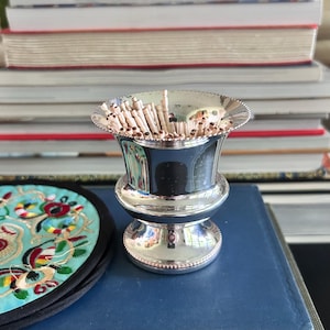 Op de afbeelding: Een zilveren tandenstokerhouder met een decoratieve kralenrand, gevuld met houten tandenstokers. De houder heeft een uitlopende bovenkant en een voetstuk. Op de achtergrond een stapel boeken en een kleurrijke geborduurde onderzetter.