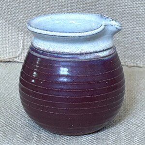 Puede incluir: Una pequeña jarra de cerámica redonda con un borde blanco y un cuerpo de color morado oscuro. El cuerpo de la jarra tiene líneas horizontales.