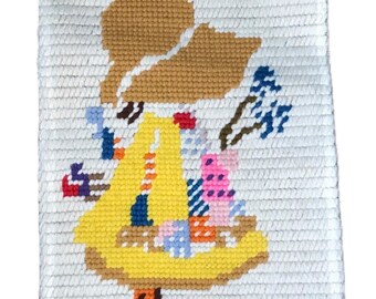 May include: A white canvas needlepoint with a colorful image of a girl wearing a yellow dress and a brown hat. The girl is carrying a basket with flowers and other items.