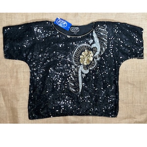 Könnte beinhalten: Schwarzes Pailletten-Oberteil mit rundem Ausschnitt und kurzen Ärmeln. Das Oberteil hat ein verziertes Blumendesign aus goldenen und silbernen Pailletten auf der Vorderseite. Ein Etikett mit dem Text "J.L.B" ist am Ausschnitt befestigt. Das Kleidungsstück ist wahrscheinlich Vintage.