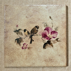 Puede incluir: Azulejo cerámico cuadrado con un diseño pintado de dos pájaros posados en una rama, una gran flor rosa y flores moradas más pequeñas. El fondo es beige texturizado.
