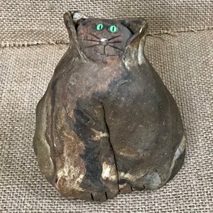 Puede incluir: Una figurita de cerámica de un gato con un esmalte marmoleado marrón y blanco. El gato tiene ojos verdes y un bigote blanco.