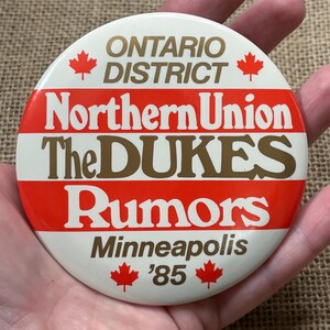 Pode incluir: Botão branco com listras vermelhas e brancas. O botão diz "Ontario District Northern Union The Dukes Rumors Minneapolis '85" com uma folha de bordo canadense de cada lado do texto.