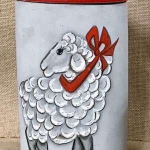 May include: White ceramic cookie jar with a red lid and a hand-painted image of a white sheep with a red bow. The sheep is facing to the left and has a black nose and eyes.