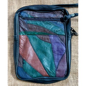 May include: A rectangular crossbody bag with a patchwork design. The bag features a black leather exterior with a colorful patchwork front in shades of green, purple, brown, and blue. It has a zippered top, a front pocket, and a shoulder strap.