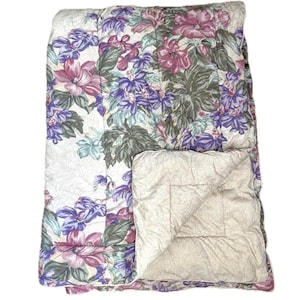 May include: A floral patterned comforter with a cream background. The comforter has a large floral design with pink, purple, and green flowers. The comforter is folded in half and the inside is visible, showing a cream-colored fabric.