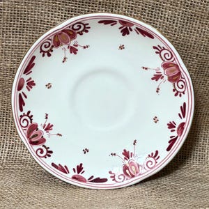 May include: A white ceramic saucer with a scalloped edge and a gold rim. The saucer is decorated with burgundy floral patterns and accents. The design includes stylized flowers and leaves, creating a vintage aesthetic. The saucer is likely part of a tea set.