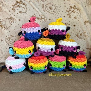 May include: A collection of nine small, round, crocheted plush toys arranged in a pyramid. Each toy features a unique color pattern, black safety eyes, pink cheeks, and a small stem on top. The text @lilithflowerart is visible at the bottom.