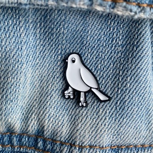 May include: White enamel pin of a bird wearing roller skates. The bird is outlined in black and has a white body. The roller skates are black with white wheels.
