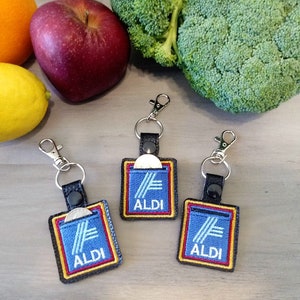 May include: Three keychains with the ALDI logo, featuring a blue background with white stripes and the word "ALDI" in white. Each keychain has a black leather strap and a silver clasp. Fruits and vegetables are in the background.