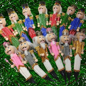 May include: A collection of nutcracker-themed pens featuring painted wooden heads with white beards, crowns or hats, and uniforms in various colors including red, green, blue, pink, silver, and gold, with white pleated fabric bases.