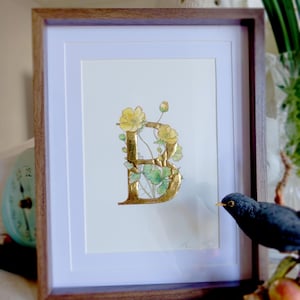 May include: A framed watercolour painting of the letter "B" with gold leaf accents. The letter is surrounded by yellow flowers and green leaves. The frame is made of wood and has a light brown finish.