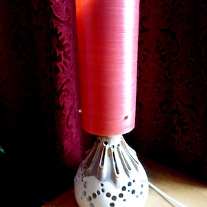 May include: A retro table lamp featuring a red cylindrical lampshade atop a decorative base. The base is white with a pattern of small holes and vertical grooves, and a tan-colored top. The lamp is plugged into a white cord.