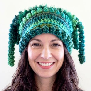 May include: A crochet beanie in shades of green featuring a textured bobble stitch pattern and long, spiralling tendrils hanging from both sides. The hat is crafted from yarn in various green tones, including emerald, lime, and forest green hues.