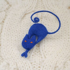 May include: A blue felt cat with orange eyes, curled up on a white knitted surface.