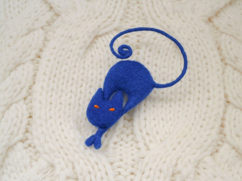 May include: A blue felt cat with orange eyes, curled up on a white knitted surface.