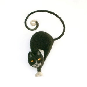 May include: A black felt cat ornament with a curled tail and orange eyes. The cat is depicted stretching, with white detailing on its face and paws. The background is plain white.