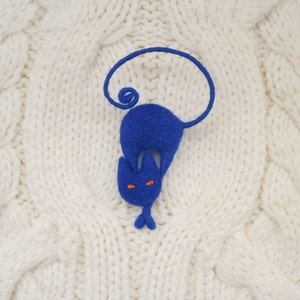 May include: A blue felt cat with orange eyes, curled up on a white knitted surface.
