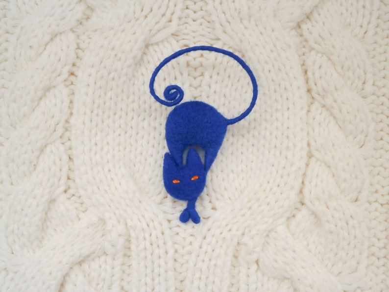 May include: A blue felt cat with orange eyes, curled up on a white knitted surface.