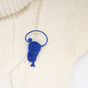 May include: A blue felt cat brooch with orange eyes, curled up on a white knitted sweater.