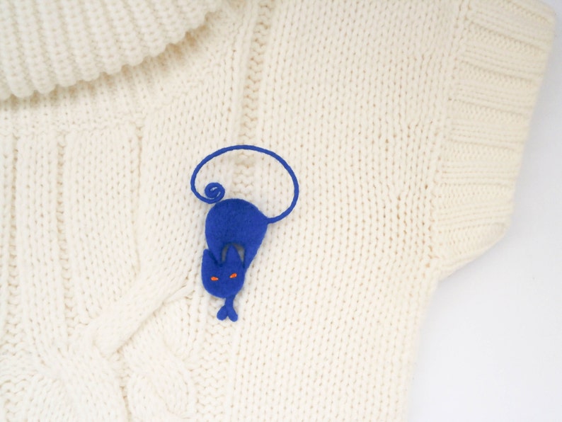 May include: A blue felt cat brooch with orange eyes, curled up on a white knitted sweater.